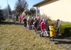 Dzieci maszerują z wiosennymi plakatami, panią Wiosną pod budynkiem przedszkola.